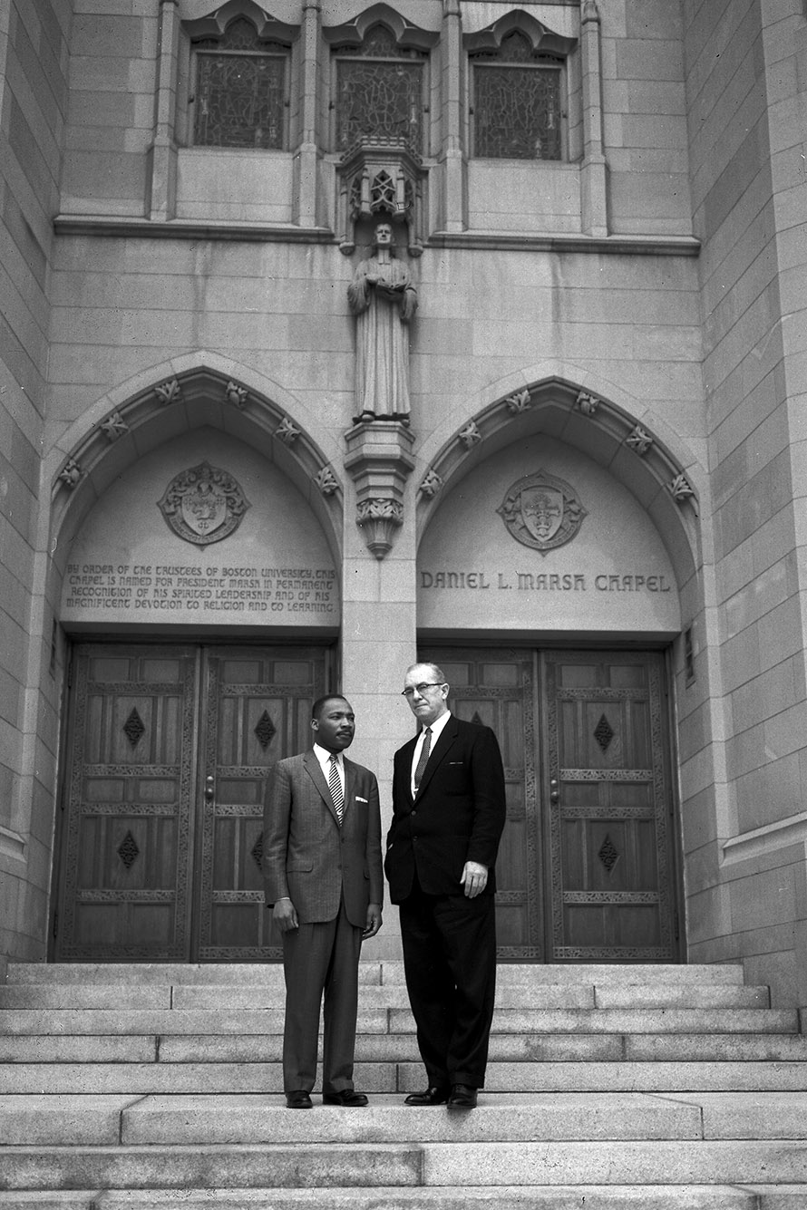 Photo: Black and white photo of Martin Luther King Jr. (left) standing next to Harold C. Case on the steps of Marsh Chapel. A Black man wearing a suit ensemble and tie stands next to a taller white man wearing a black suit ensemble and tie. The both stand on steps in front of large church doors.