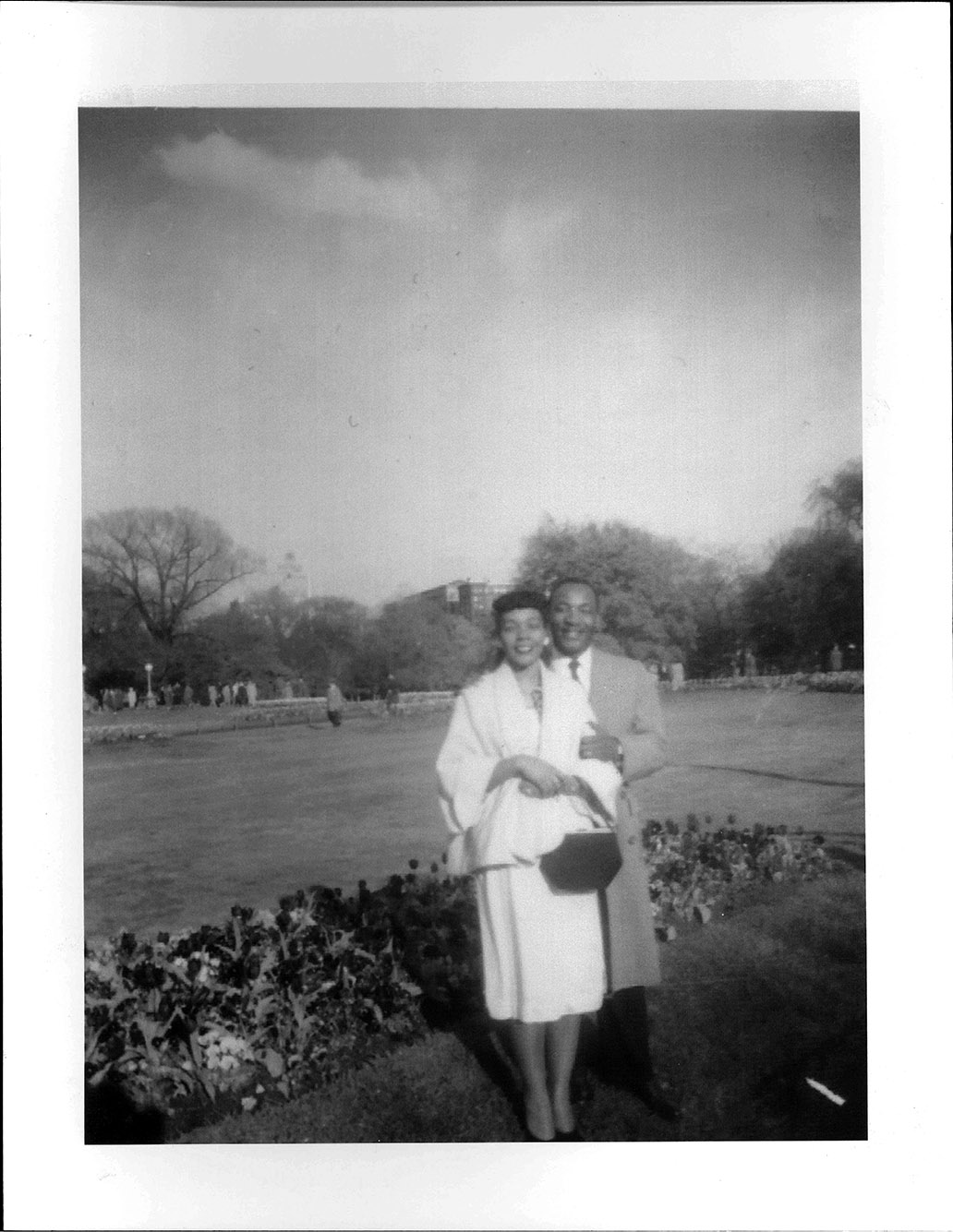 Photo: black and white photo of Martin Luther King Jr. and his future wife Coretta Scott, posing in a scenic park. A Black mean wearing a suit, tie, and coat, smiles and poses behind a Black woman wearing a white jacket and skirt.