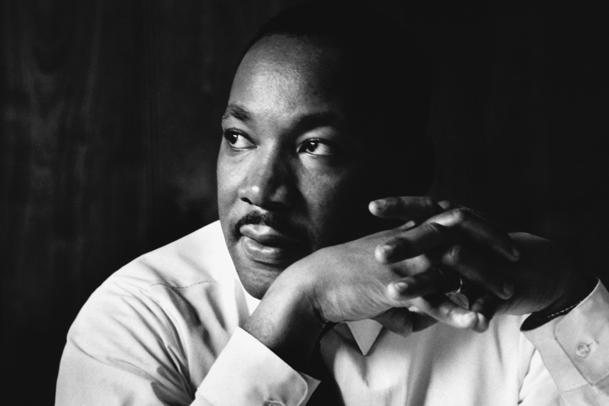 Photo: Black and white photo of Martin Luther King Jr., a Black man wearing a long-sleeved white collared shirt and tie, looking to the left pensively. He clasps his hands in front of his face.