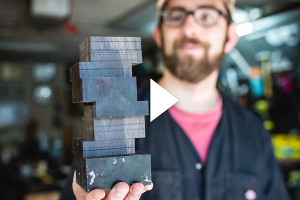 Photo: Micah Sieber, a white man with brown beard and mustache and wearing a tan cap, red-pink shirt, black collared shirt, and black pants, smiles and holds up a small black, model building. The building is in the shape of the BU CDS building, a Jenga-like building with smooth sides. A white triangle play button is overlaid in the center.