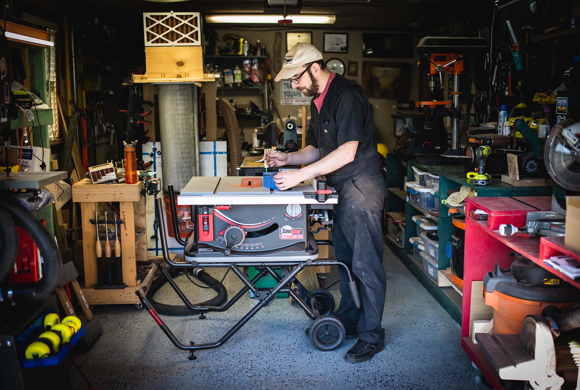 Photo: Micah Sieber, a white man with brown beard and mustache and wearing a tan cap, red-pink shirt, black collared shirt, and black pants, bends over a woodworking table and works on a piece in his home wood working space.