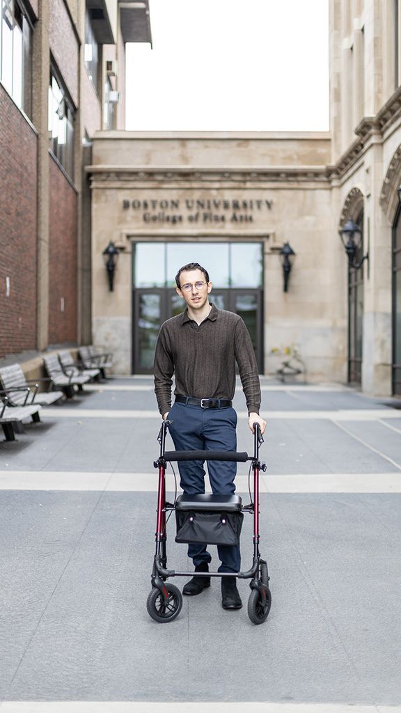 Photo: A young white man wearing glasses, a brown, long sleeved collared shirt and navy blue slacks, stands and poses with the aid of a mobility device. He poses in front of BU's College of Fine Arts.