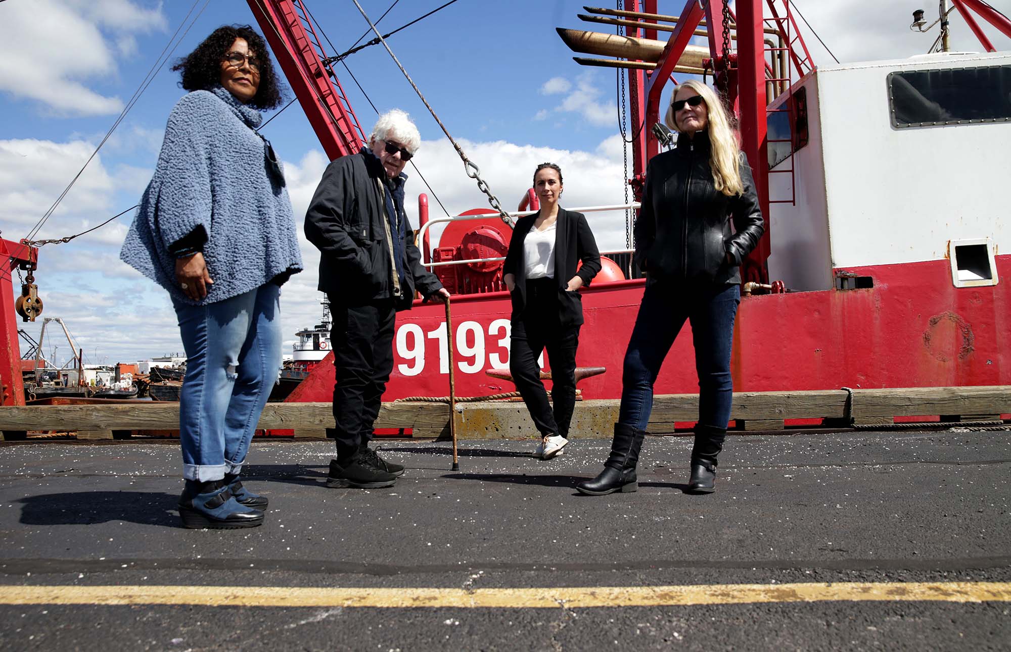 Photo: Four people in business casual attire stand and pose in front of a large red ship docked in a port on a sunny day. From left, Jeanne Costa, Ken Hartnett, Daniela Melo and Barbara Roessner pose for a photo.