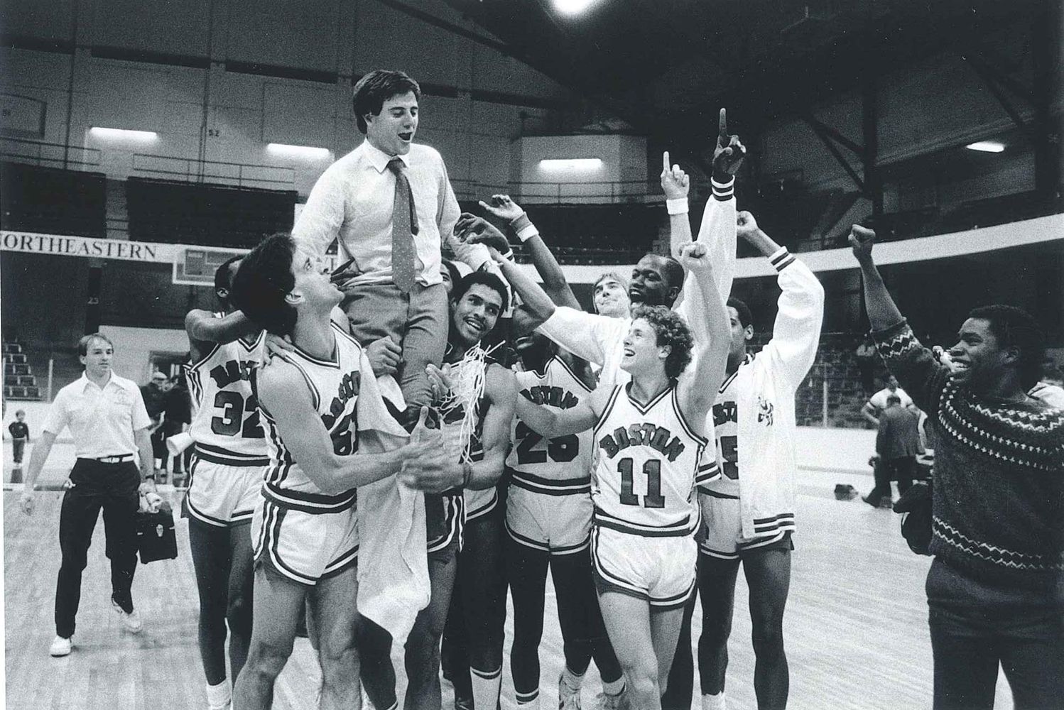 Photo: A black and white photo of Rick Pitino celebrating with the BU men's basketball time he coached. Members of the team hoist him on their shoulders as they group up and cheer on the basketball court.