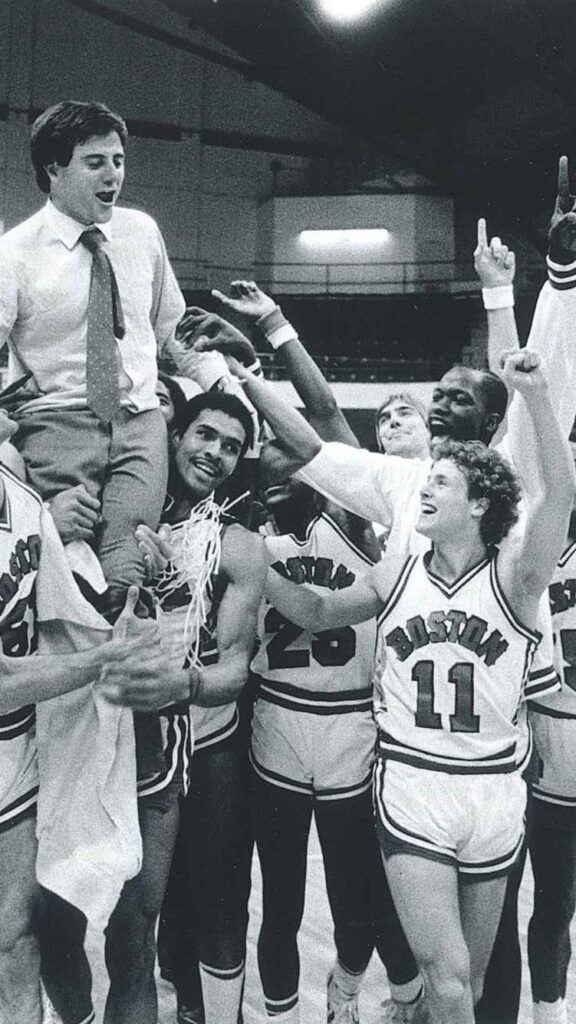 Photo: A black and white photo of Rick Pitino celebrating with the BU men's basketball time he coached. Members of the team hoist him on their shoulders as they group up and cheer on the basketball court.
