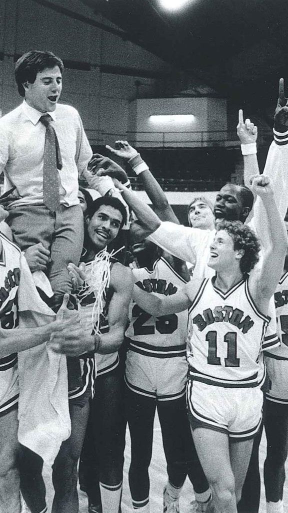 Photo: A black and white photo of Rick Pitino celebrating with the BU men's basketball time he coached. Members of the team hoist him on their shoulders as they group up and cheer on the basketball court.