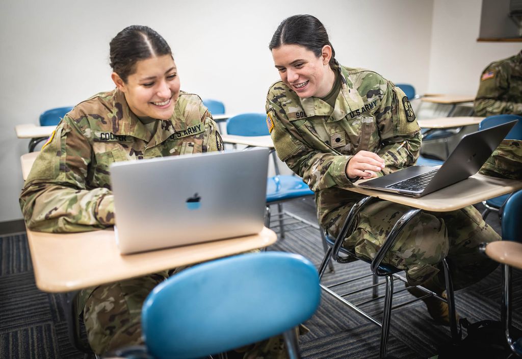 Photo: Two young women wearing Army ROTC uniforms sit at desks next to each other and smiles. The student on the right looks at something the student on the left is sharing on her grey laptop.