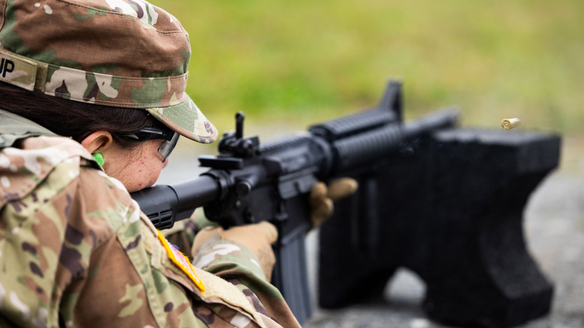 Photo: A young woman wearing ROTC army fatigue gear is shown lying on the ground looking into the eye piece of a black rifle as she aims to shoot at a gun range.