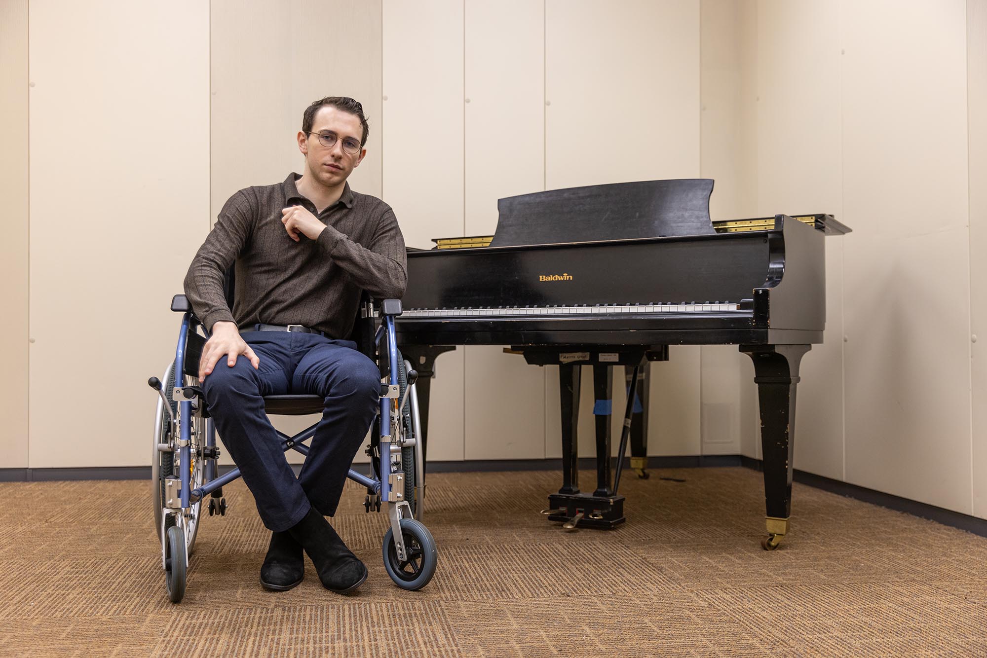 Photo: A young white man wearing glasses, a brown, long sleeved collared shirt and navy blue slacks, sits in a wheelchair and poses for the photo next to a large piano.