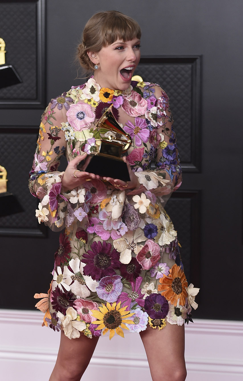 Photo: Taylor Swift, a white woman with dirty blonde hair and wearing an elaborate dressed made of fake flowers, shows a shocked reaction as she golds a Grammy award in her hands.