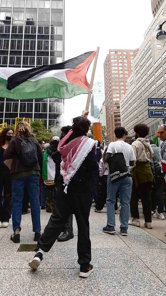 Photo: A group of protestors are shown standing in front of the Consulate General of Israel along Second Avenue, New York, NY. A person in the center holds a large Palestinian flag above the crowd.