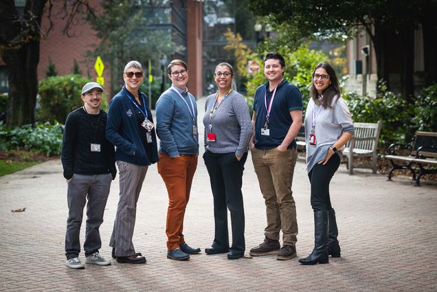 Photo: A group of individuals from the Boston Medical Center’s, GenderCare team pose with their hands in their pockets, smiling at the camera. There are six of them, circling around to make a half-moon. They stand outside, on a bright day.