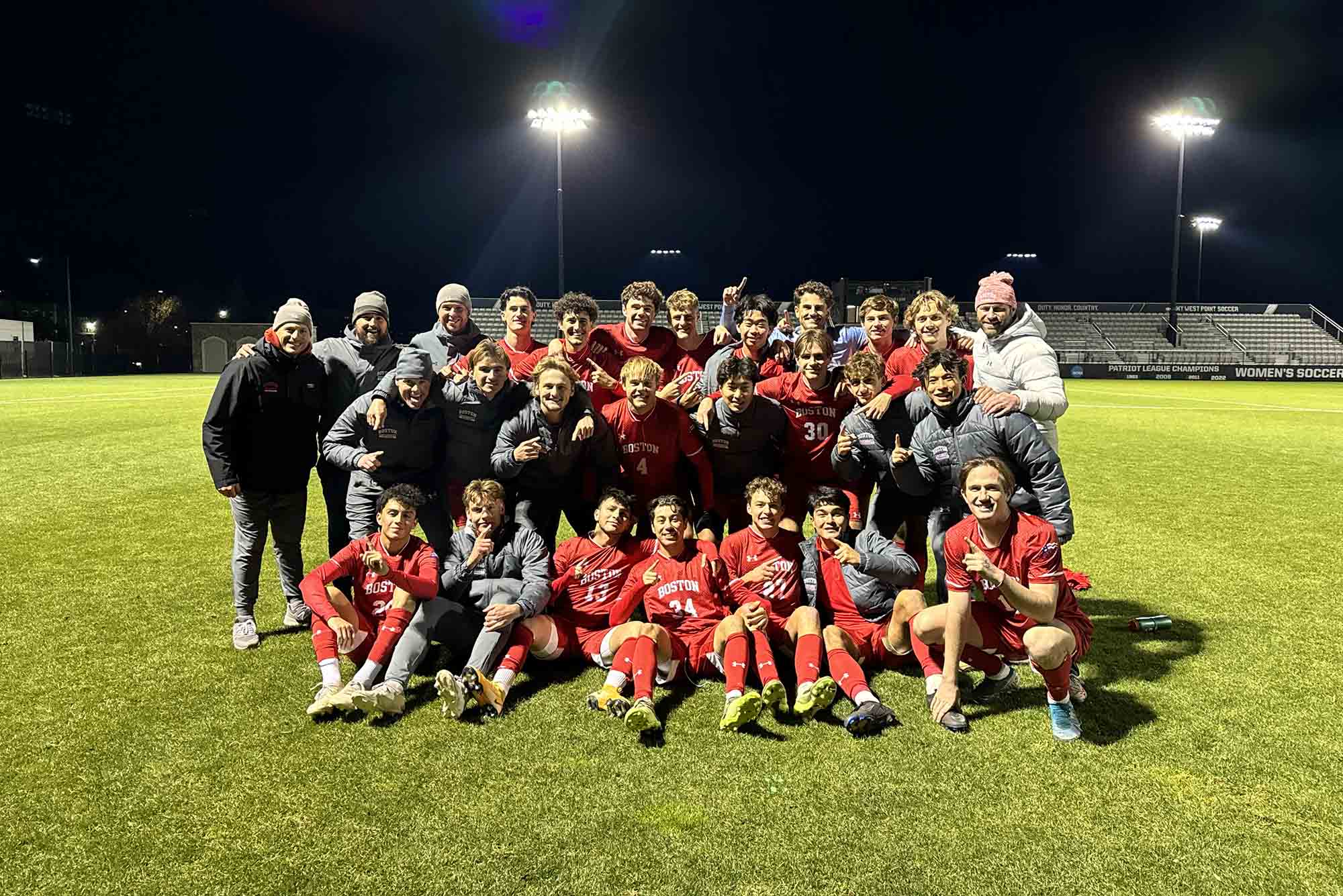 Photo: A group photo of the BU men's soccer team in their red uniforms. They are on a soccer field, smiling with their coaches.
