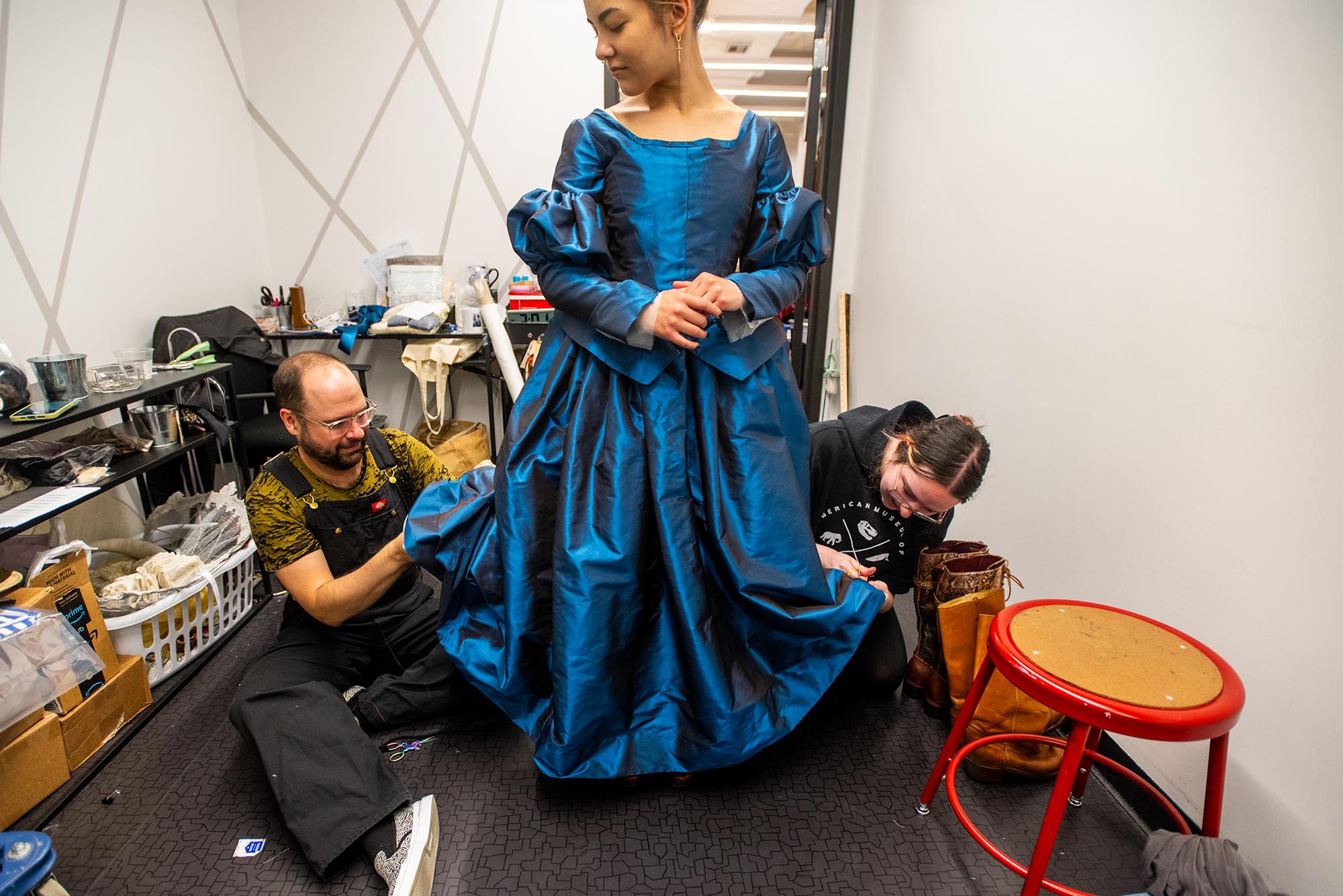 Photo: A women stands in a regal blue dress as the costume designers make amends to the garment. She looks down to watch them work.