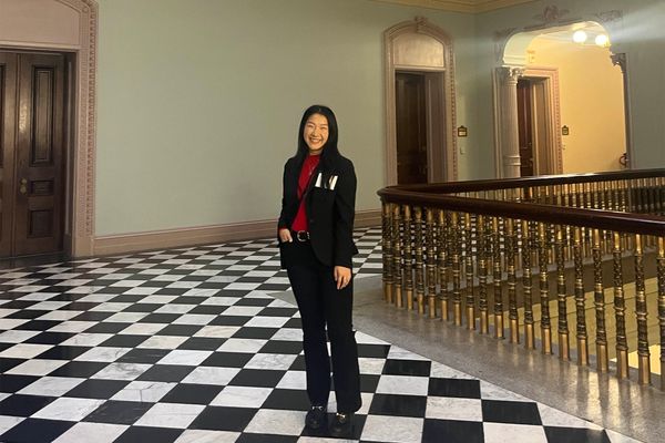 Photo: A smiling woman in a suit and blazer stands in a checkered floor in an ornate room near a wooden carved railing