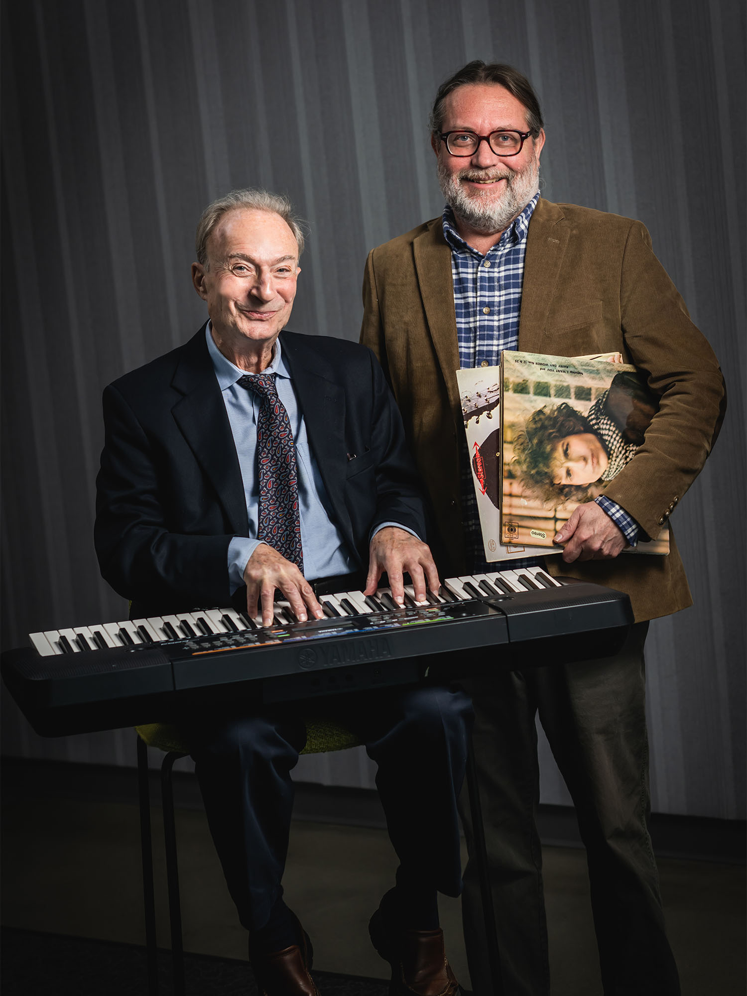 Photo: Two men in suits sitting in front of a piano with smiles on their faces. One is holding a Bob Dylan LP