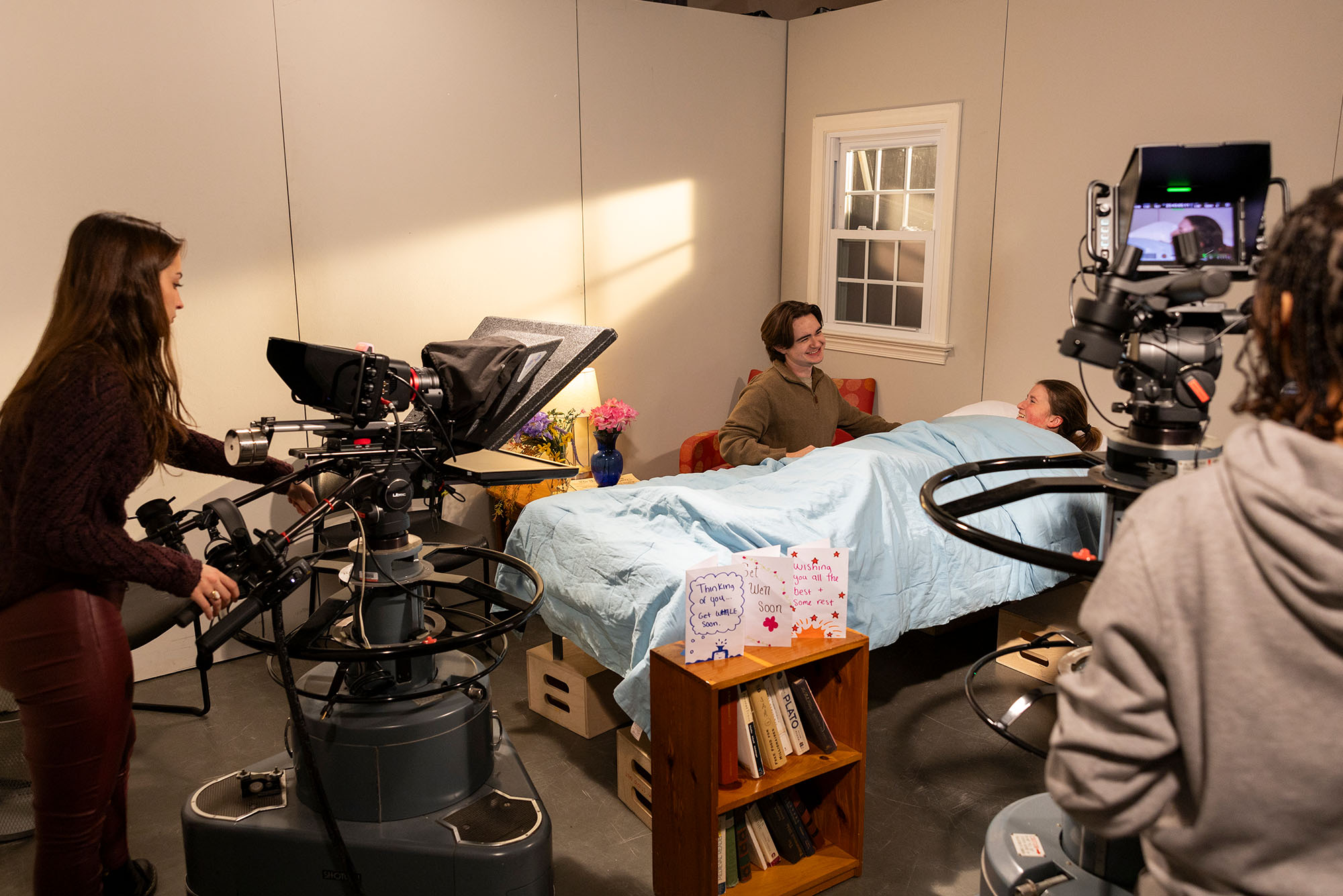 Photo: A group of college students in a sound stage, producing a TV medical drama. There is a student laying in a hospital bed and someone sitting at the beside singing a musical number, while cameras roll.
