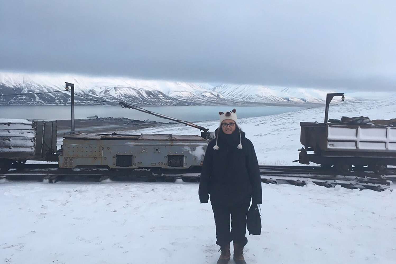 Photo: Adriana Craciun, a CAS professor of English, outside Mine 3, a shuttered coal mine near Longyearbyen in Svalbard. A white woman bundled up in black winter gear stands and poses in front of an old coal railway in a snowy landscape. Snowy mountains can be seen in the distance.