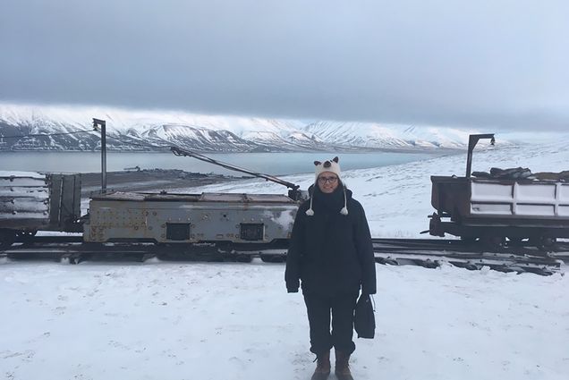 Photo: Adriana Craciun, a CAS professor of English, outside Mine 3, a shuttered coal mine near Longyearbyen in Svalbard. A white woman bundled up in black winter gear stands and poses in front of an old coal railway in a snowy landscape. Snowy mountains can be seen in the distance.