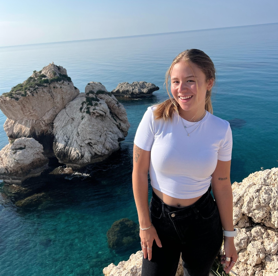 Boston University student Pamela Arjorna. She is standing in front of the blue ocean with cliff rocks behind her. She is wearing black jeans and white plain crop top. She has her blonde hair pulled back and she is smiling at the camera. 