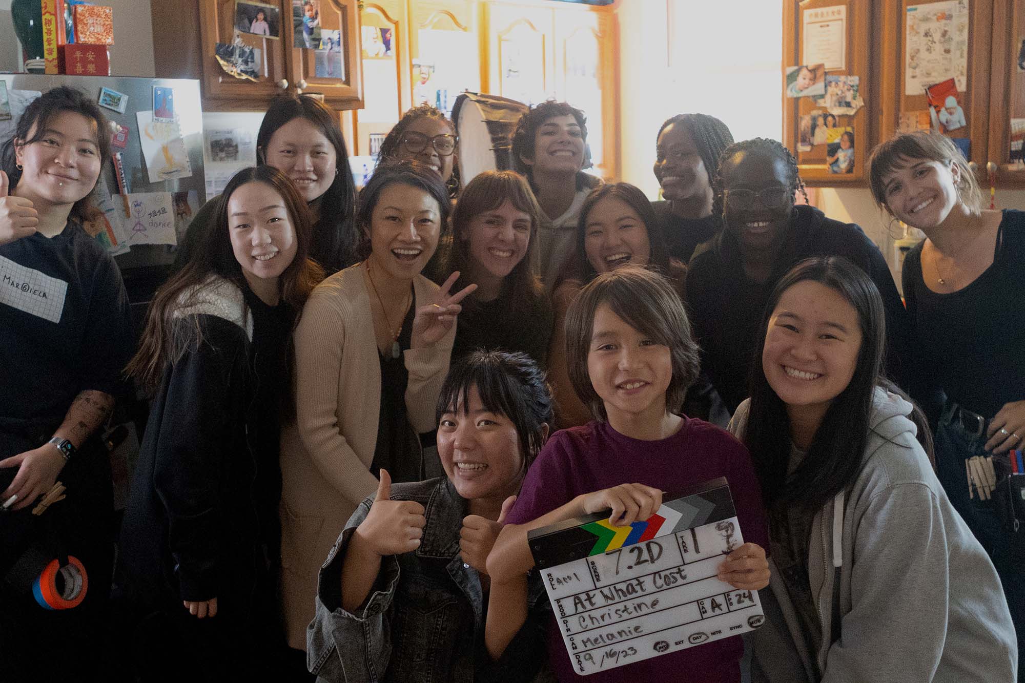 Photo: A large group of people gather and pose together in a living room film set. A person front and center of the group holds a marquee that reads "At What Cost".