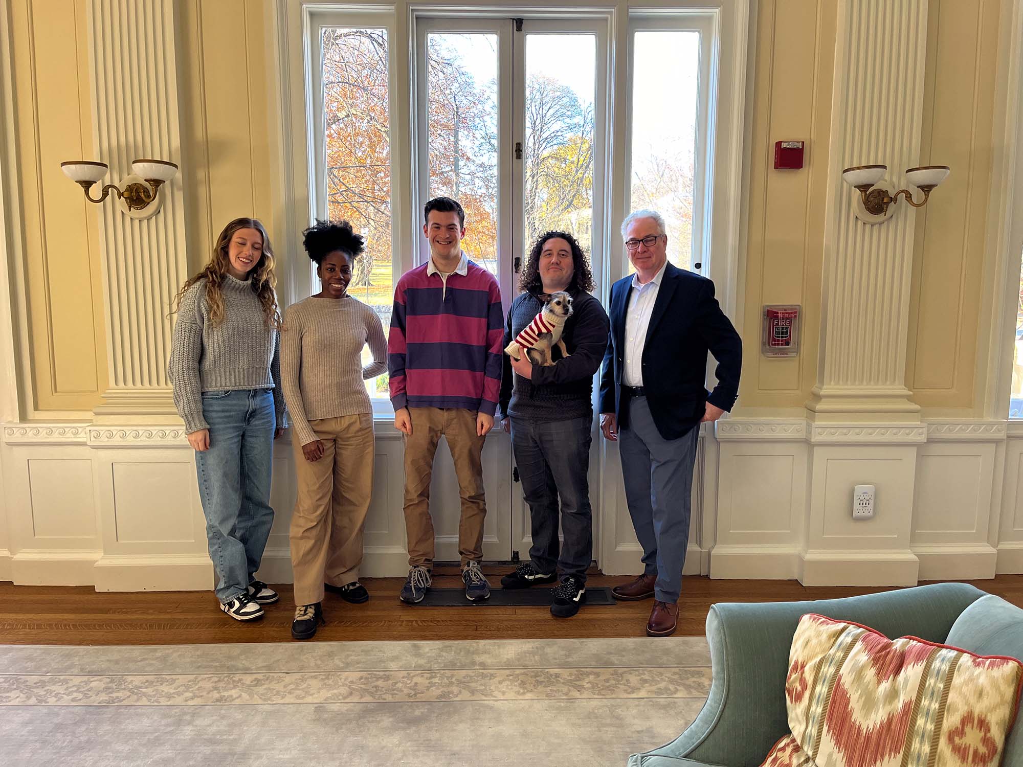 Photo: A diverse group of people pose in front of large window and appear shadowy as a result. From left to right: a young white woman wearing a grey sweater and jeans, a young Black woman wearing an oatmeal-colored sweater and khaki pants, a young white man wearing a pink and blue striped collared shirt, a white man with curly hair holding a small dog wearing a holiday sweater, and an older white man wearing glasses, a light blue collared shirt, navy blue blazer, and jeans.