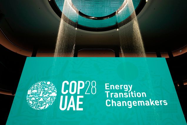 Photo: A large teal sign reads "COP28 UAE: Energy Transition Changemakers". Streams of lit up water streams are shown in front of the bright sign.