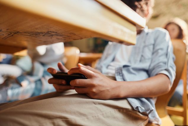 Photo: A zoomed in photo shows a young student discretely using their cell phone under their desk as they sit in the classroom.