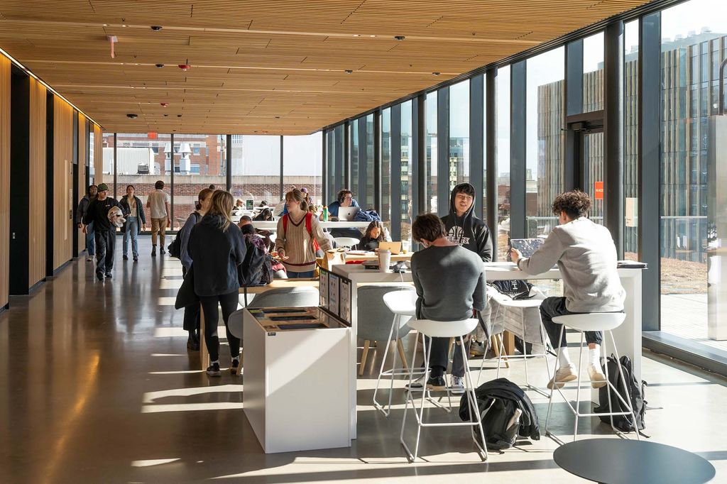 Photo: Members of the BU community study in the 5th floor lounge inside of the BU Faculty of Computing & Data Sciences (CDS) Building. People mingle and sit in a large open, brightly lit space. They sit in white bar chairs at various large white tabletops and mingle, study, and nibble on snacks.