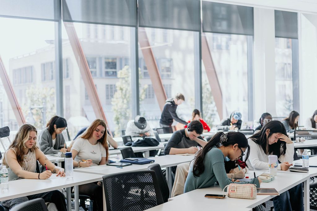 Photo: A group of students are shown sitting at white tables in an open space classroom. Behind them, large floor to ceiling windows let in tons of natural light.