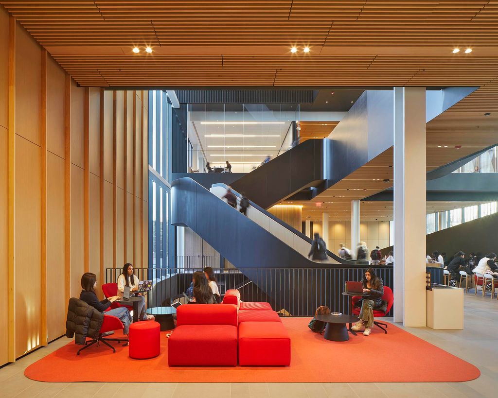 Photo: Interior of Boston University's Center for Computing & Data Sciences. Interior shows high ceilings featuring wood and black accents. Two large opens spaces with bright red modular furniture is shown as student sit, mingle, study, and walk in and around the space.