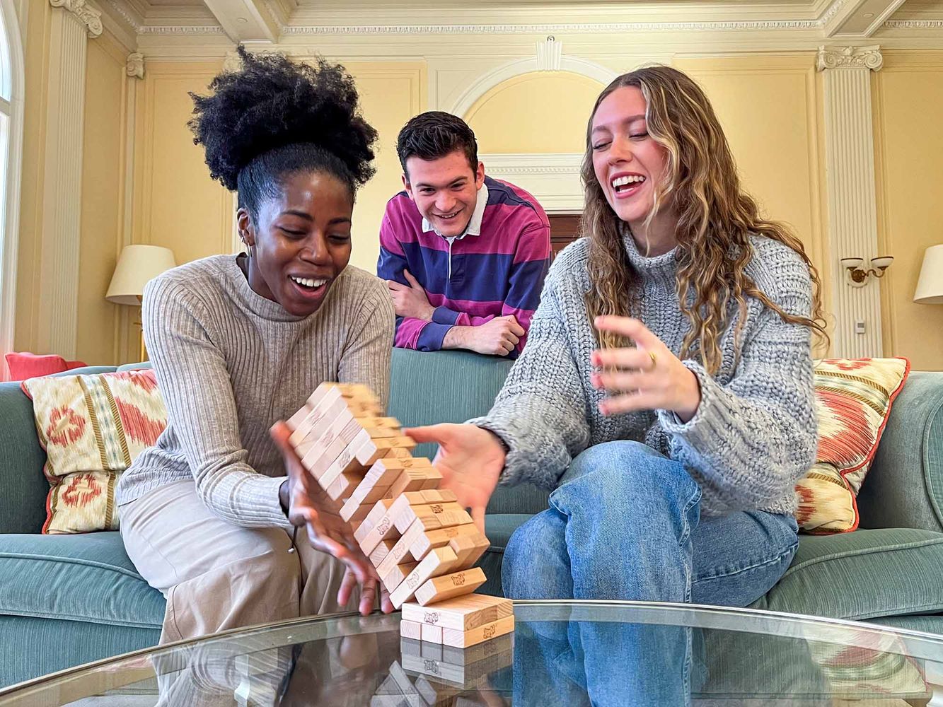 Photo: A Black woman with hair tied up in an afro puff and wearing a an oatmeal-covered sweater and tan pants sites to the left of a young white woman with long brown hair and wearing a grey sweater and jeans. They both sit on a muted green couch. Leaning on the back of the couch and watching the game is a young white man wearing a pink and blue striped collared shirt. All three look happy and surprised as the Jenga game on the table in front of them collapses.