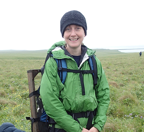 Photo: Catherine West, a smiling white woman wearing a black beanie cap, green jacket, and large backpack smiles and poses in a green field on a cloudy day.