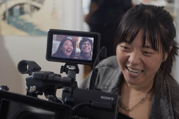Photo: A young Asian woman is shown filming using a large black film camera. The small display on the camera shows two young adults in the midst of being filmed for a scene.
