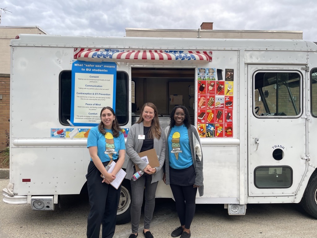 This is a photo from the Cones for Consent event from summer 2023 at Boston University's Student Orientation. There are three individuals standing in front of an ice cream truck. On the left and right, the women are in blue t-shirts that have ice cream cones on them. The person in the middle is wearing a black shirt and grey cardigan, holding up a clipboard. 