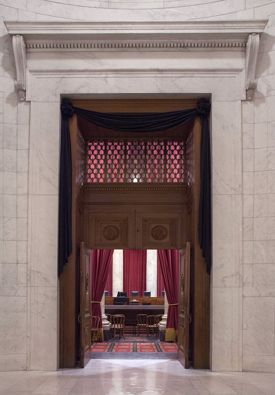 Photo: The Supreme Court courtroom doors are shown draped in black. A large wooden doorway is shown with doors open. Inside the doors the Supreme court bench can be seen along with several seating areas. A large black cloth is draped over the top and top sides of the door.