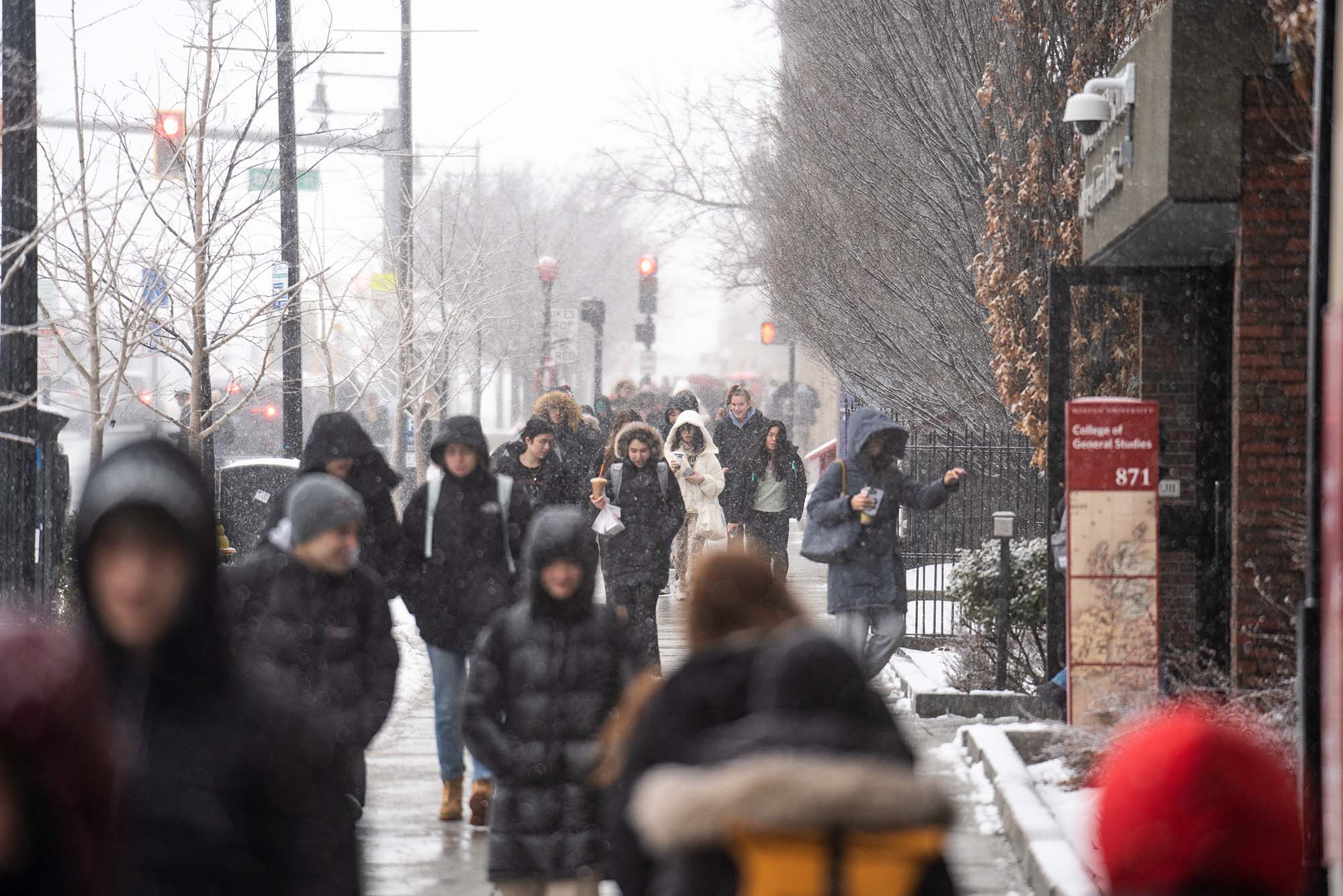 Photo: BU community members endure the elements while walking down the street next to the CGS building. Various people bundled up in winter gear are shown walking down the sidewalk on a snowy day.