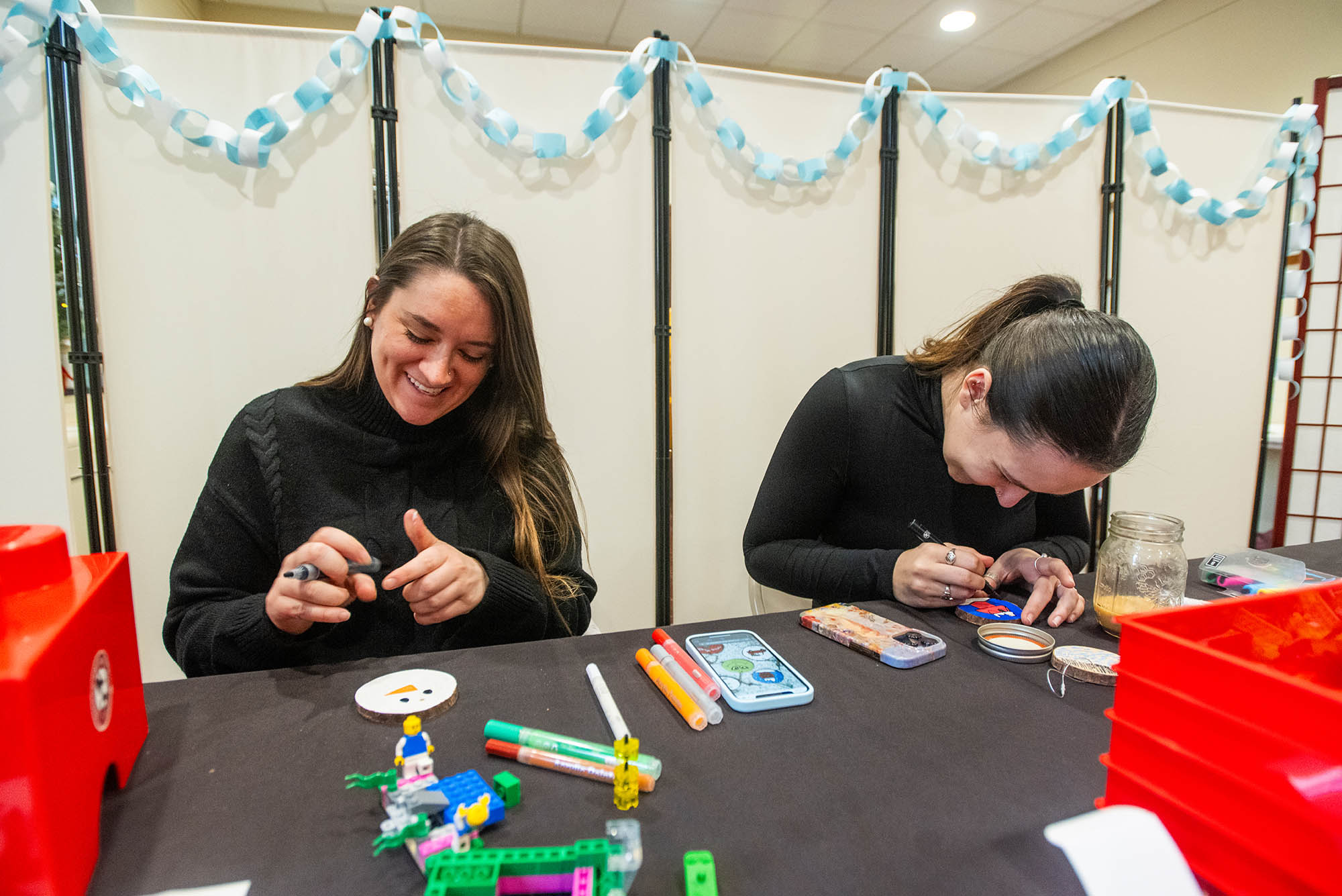 Photo: Two young women wearing black tops sit at a black table and paint flat, round ornaments in front of them with paint markers. The women on the left smiles as her ornament takes the appearance of a snowman with a carrot nose.