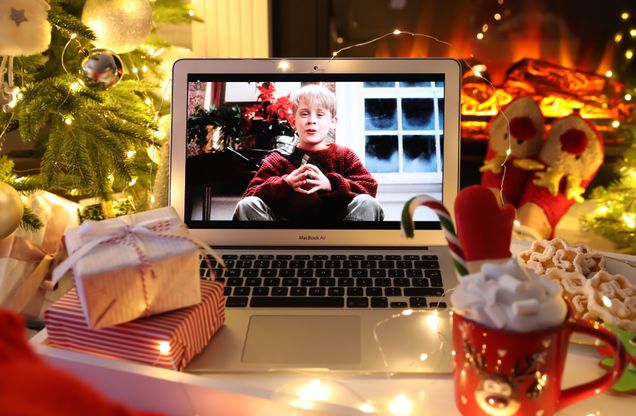 This is a holiday themed stock photo from Canva. There is holiday scenery in the background, with a tree, presents, a hot cocoa, and a laptop with the home alone movie on the laptop. There are yellow holiday lights strung up around the laptop.