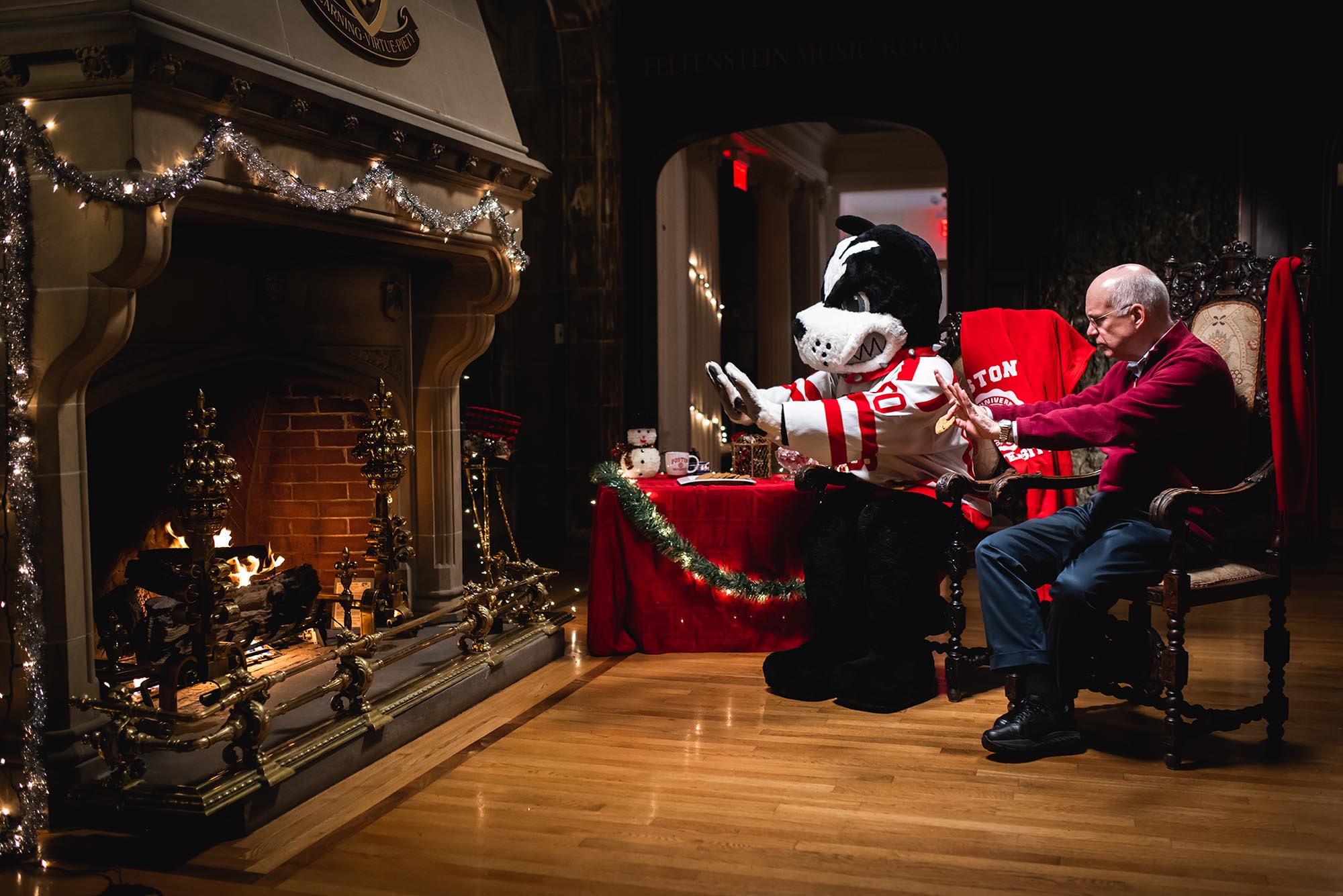 Photo: President Ken Freeman, an older, balding white man wearing glasses, a red holiday sweater, and jeans sits next to a lit fireplace with Rhett, a Boston Terrier school mascot wearing a red and white Bu Hockey jersey. They both sit and splay their arms out towards the lit fireplace to their left.