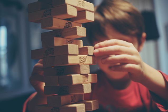 Photo: Zoomed in photo of a younger child playing Jenga. The child's face is obscured by the precarious Jenga tower.