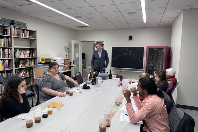 Photo: Zachary Fuller (center) shares his final project with class members and the broader Gastronomy Program at BU. A white man wearing glasses, a pink shirt, and blue cardigan listens as someone seated at a long table asks him a question. Five other people also sit at the table as various clear cups filled with various beverages are lined up in front of them.