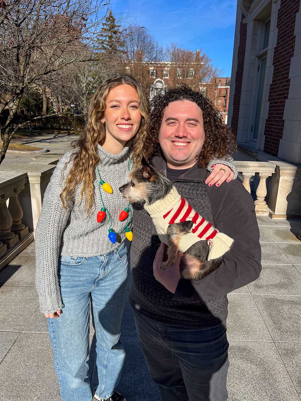 Photo: Two people stand and pose outside for a photo. On the left, a young white woman wearing a grey sweater, Christmas light necklace, and jeans poses with right arm over the shoulders of a white man with curly hair and wearing a black sweater and pants and holding a small dog wearing a holiday sweater. They both squint towards the camera and smile.