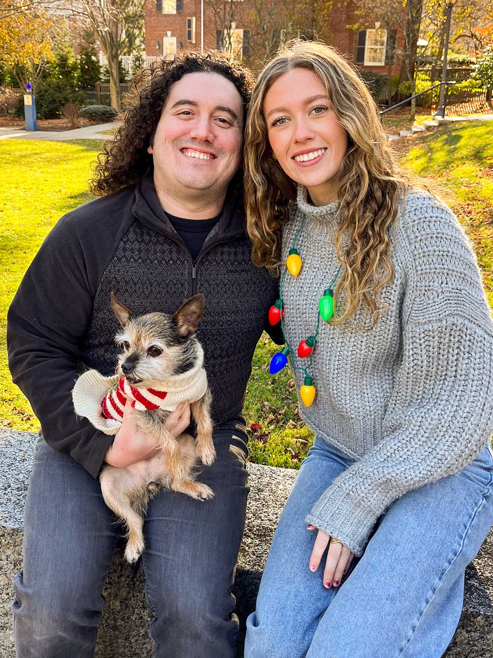 Photo: Two people stand and pose outside for a photo. On the left, a white man with curly hair and wearing a black sweater and pants and holding a small dog wearing a holiday sweater poses with right arm over the shoulders of a young white woman wearing a grey sweater, Christmas light necklace, and jeans. They both look towards the camera and smile.