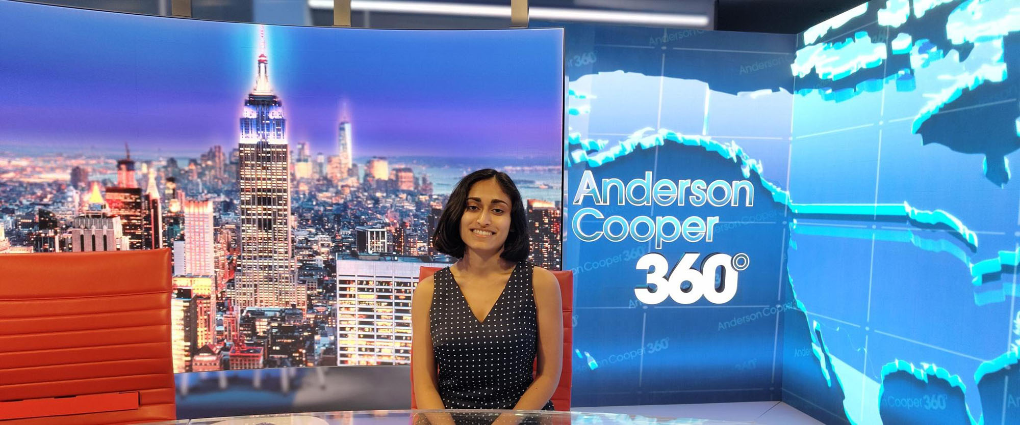 Photo: Madhri Yehiya, a young South Asian woman with short black hair and wearing a black blouse sits at a news desk in the Anderson Cooper 360 Studio. The large screen behind her displays the words "Anderson Cooper 360".