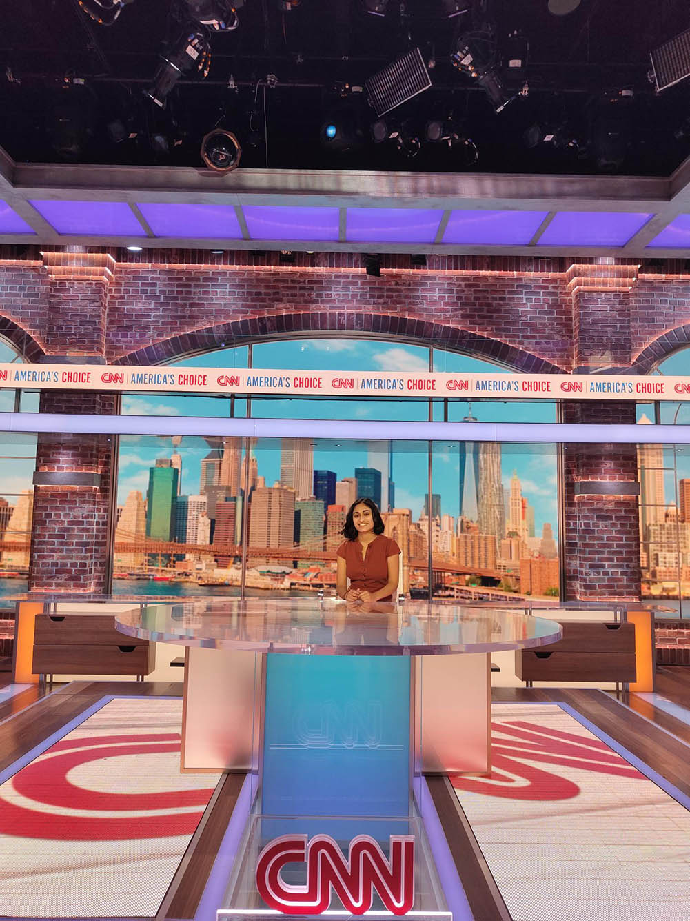 Photo: Madhri Yehiya, a young South Asian woman with short black hair and wearing a dark red blouse sits at a news desk in the CNN studios.