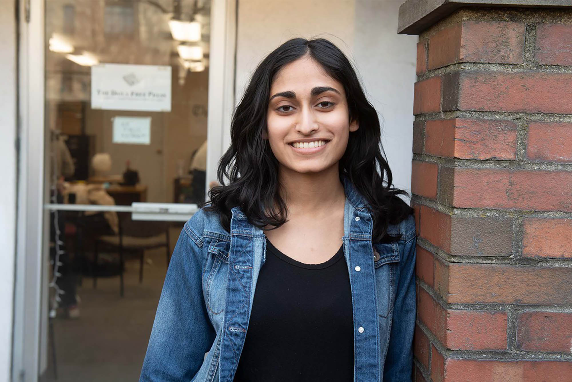 Photo: Madhri Yehiya, a young South Asian woman with curled black hair and wearing a black shirt and jean jacket smiles and poses outside a clear door of a brick building.