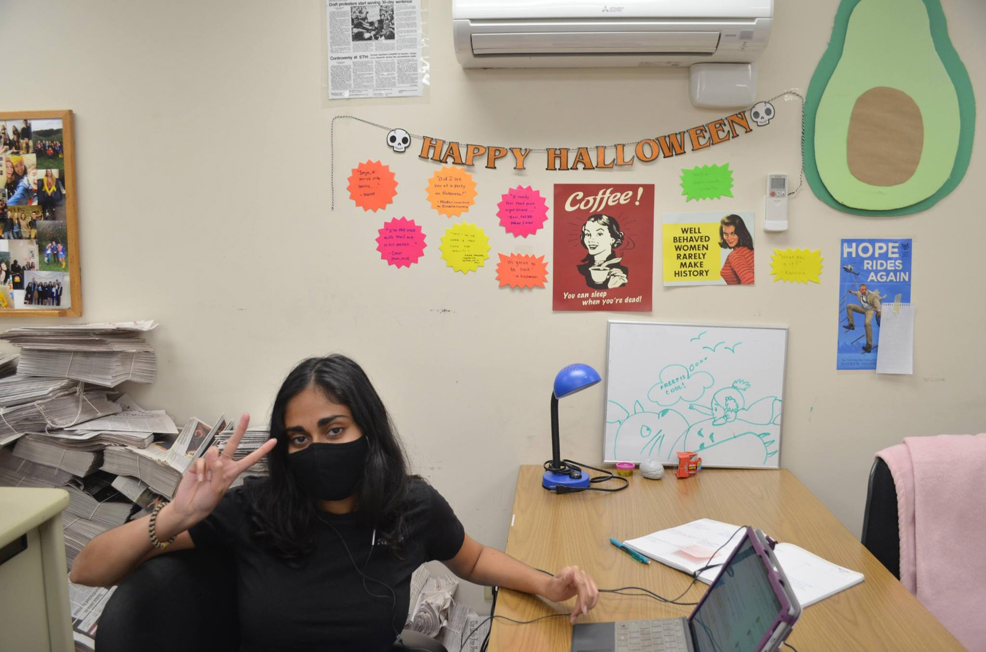 Photo: Madhri Yehiya, a young South Asian woman wearing a black mask and black shirt, looks tot he camera and poses as she sits at a desk with small laptop open in front of her. Stray papers can be seen around her.