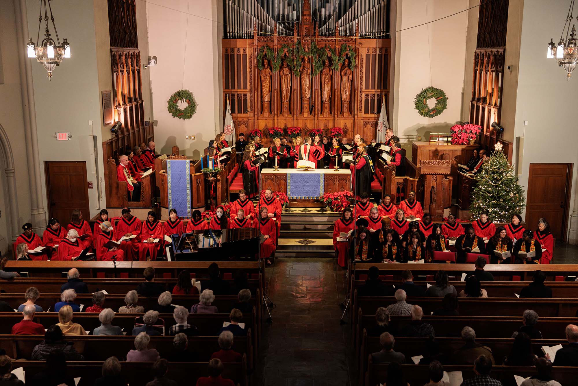 Photo: 50th Annual Service of Lessons and Carols on December 8, 2023 at Boston University's Marsh Chapel is shown. Wide shot of the service shows the carolers dressed in red robes and standing and singing with black librettos open. Pews in front of them are filled with people watching.