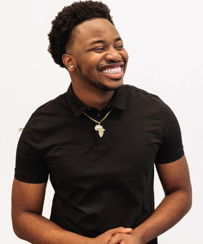 This is a photo of BU student, Nsikan Umoh (CAS'24). He is smiling and looking off to the right. He is wearing a black polo shirt with a gold chain around his neck. The photo is taken in front of a plain white back drop. 
